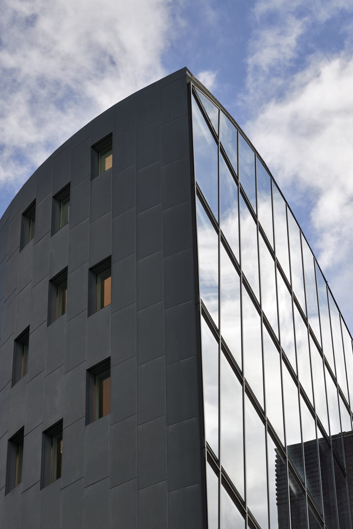 Gebäude mit grauer Fassade und Fenstern, die den Himmel und Wolken spiegeln, vor blauem Himmel.