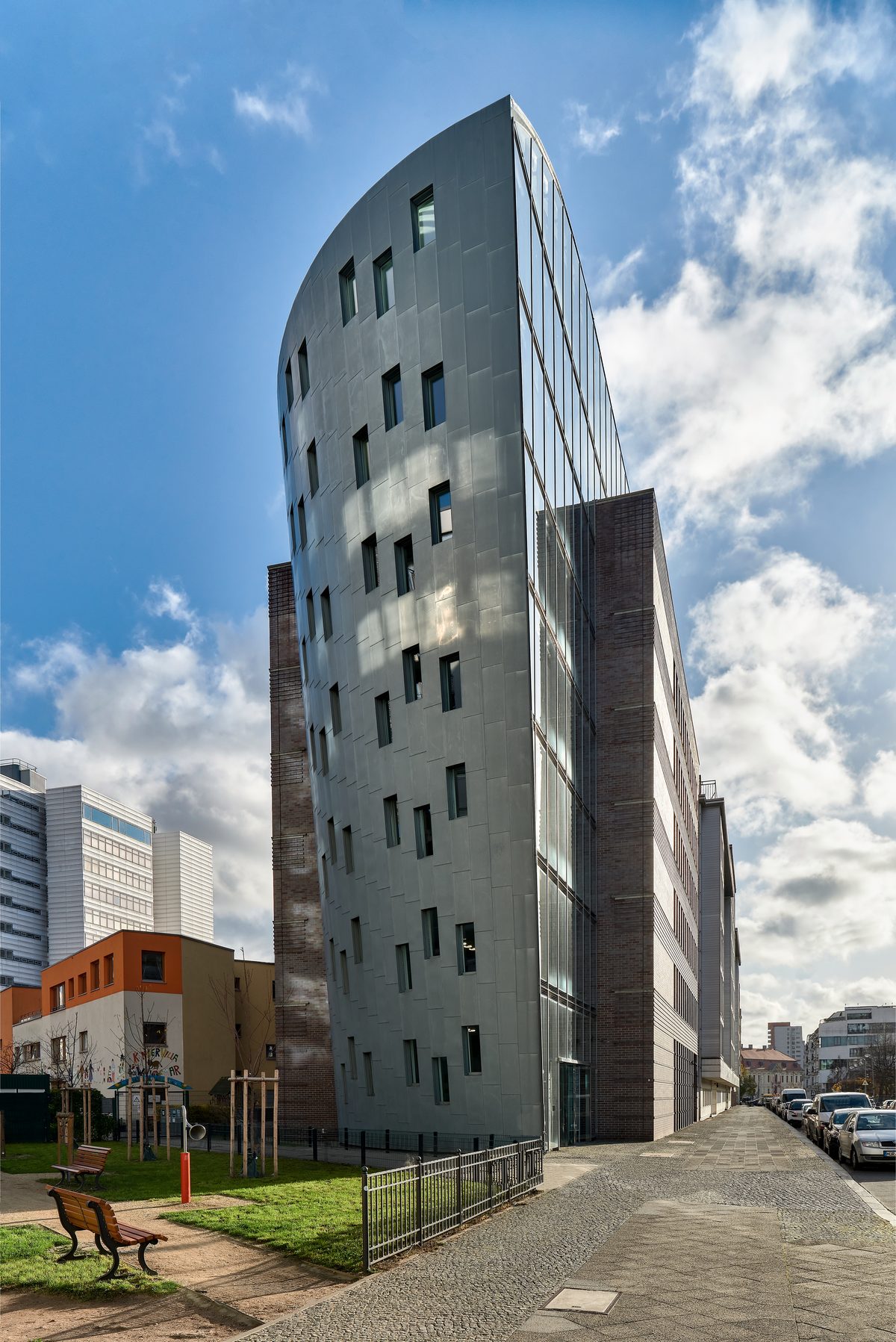 Gebäude mit geschwungener Metallfassade und Glasfront, daneben ein Bürgersteig mit parkenden Autos und eine Grünfläche.