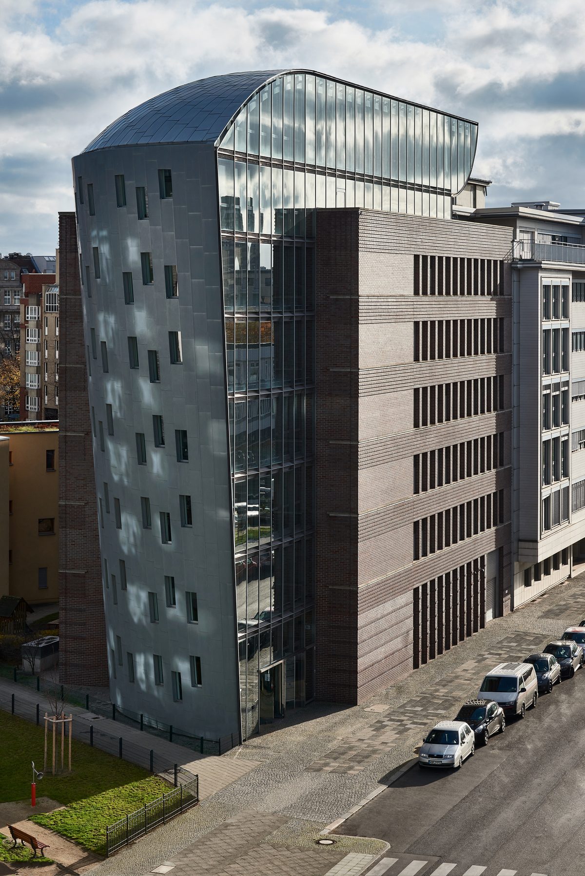 Gebäude mit geschwungener Glasfassade und Ziegelmauerwerk, davor eine Straße mit parkenden Autos und ein Grünstreifen.