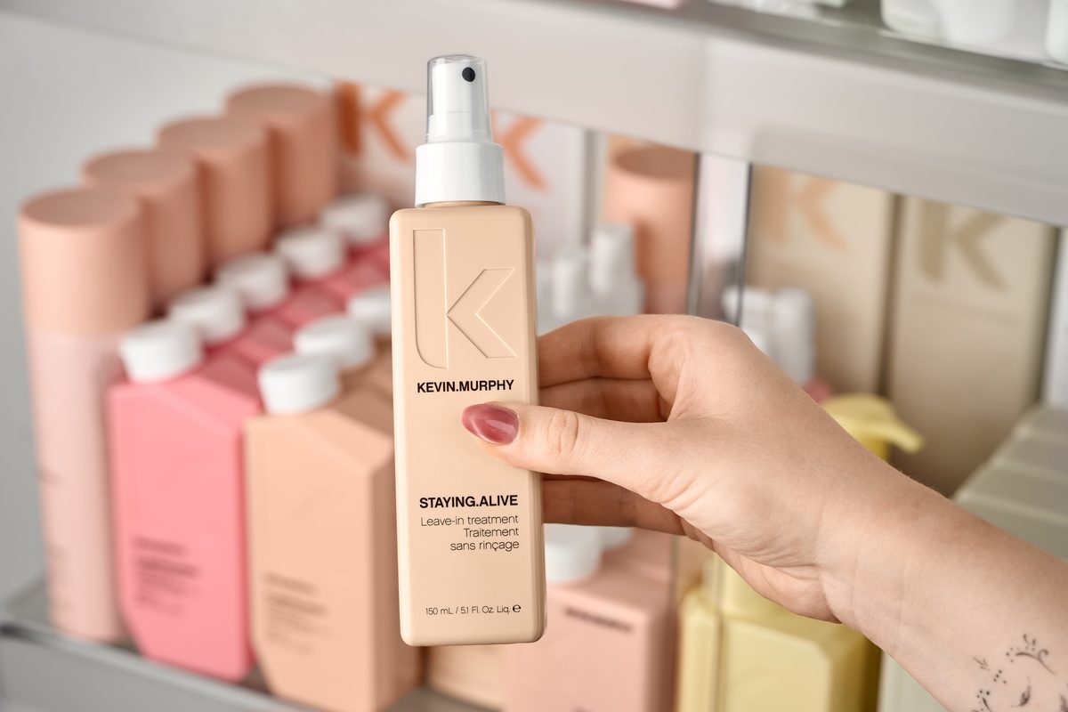 Eine Hand hält eine beige Sprühflasche mit einer Aufschrift vor einem Regal mit weiteren Flaschen.