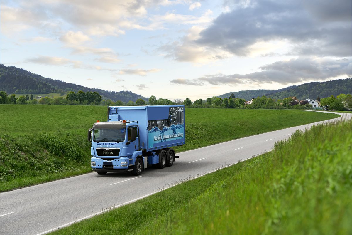 Ein blauer LKW mit einer Aufschrift fährt auf einer Landstraße durch eine grüne Hügellandschaft.