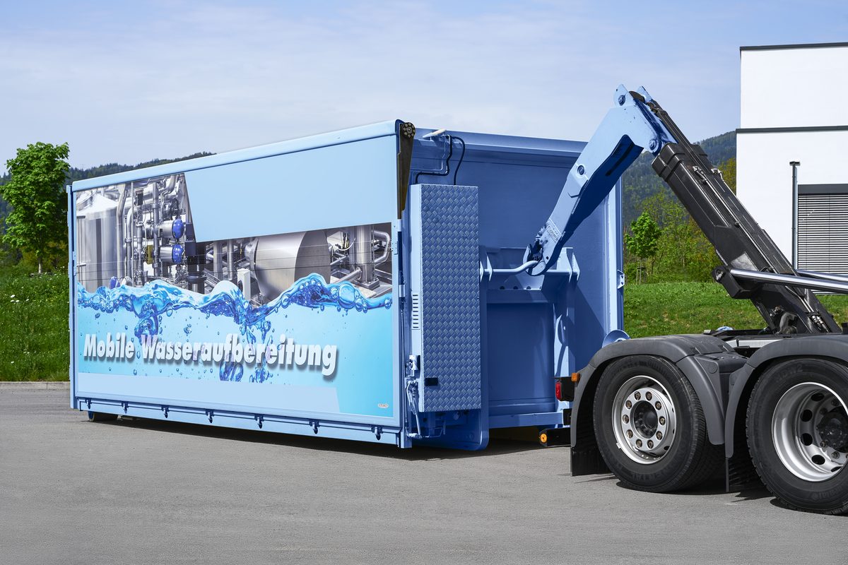 Ein blauer Container mit einer Aufschrift wird von einem LKW-Hakenlift abgesetzt.
