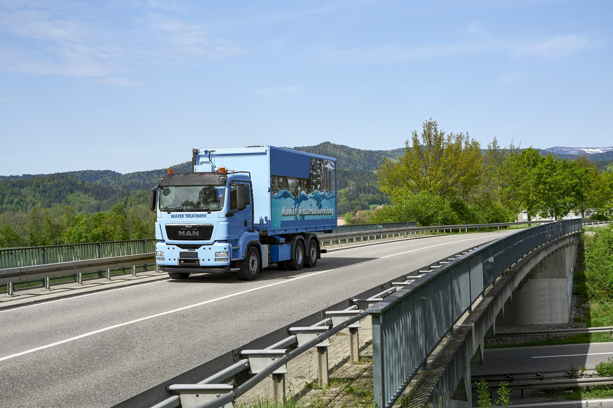 Ein blauer MAN-LKW mit einer Aufschrift fährt auf einer Brücke durch eine grüne Landschaft.