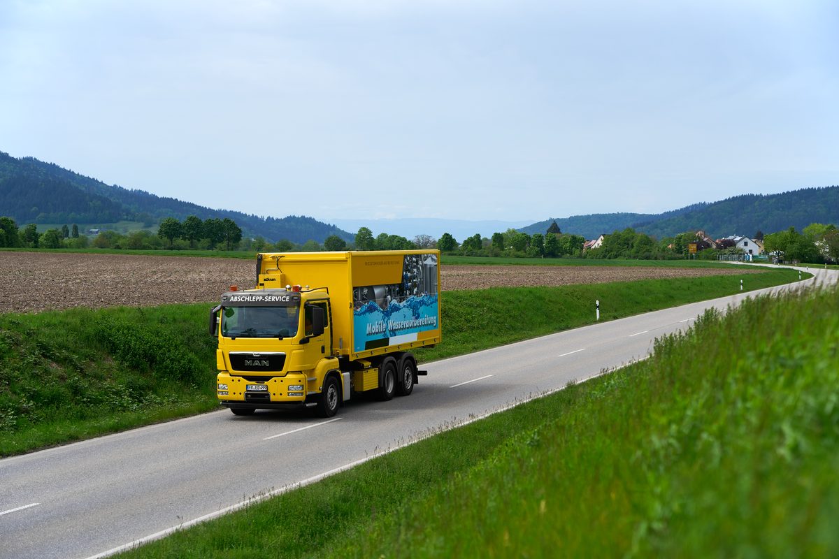 Ein gelber LKW mit einer Aufschrift fährt auf einer Landstraße durch eine hügelige Landschaft.