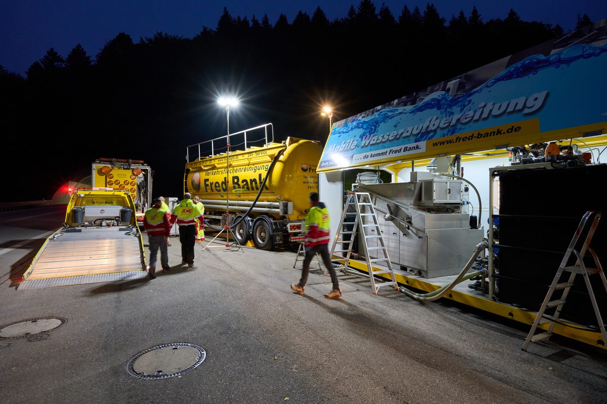 Drei Arbeiter stehen nachts neben einem gelben Tanklastwagen und einer mobilen Wasseraufbereitungsanlage auf einer Straße.