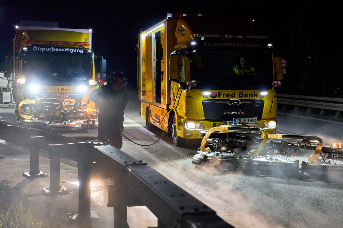 Zwei gelbe Lastwagen mit Ölspurbeseitigungsgeräten reinigen nachts eine Fahrbahn, ein Arbeiter steht daneben.