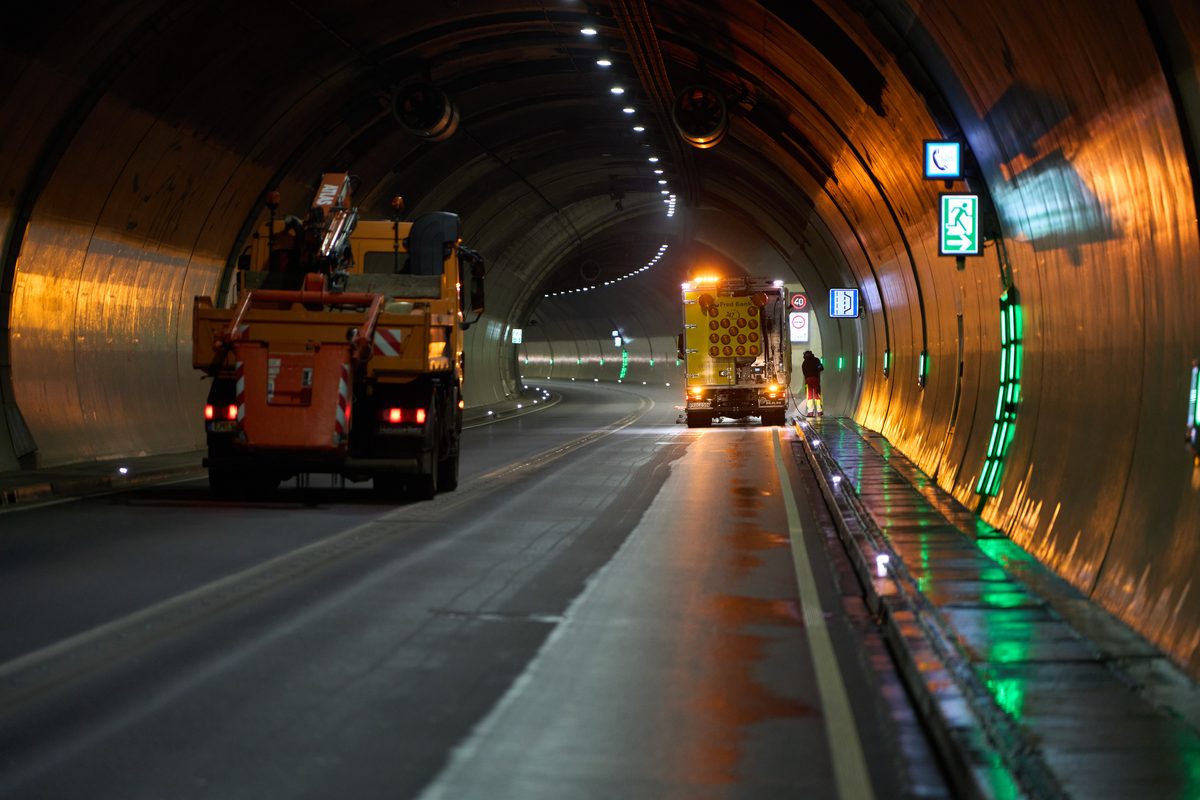 Zwei orangefarbene Wartungsfahrzeuge stehen in einem beleuchteten Tunnel, ein Arbeiter steht neben dem hinteren Fahrzeug.