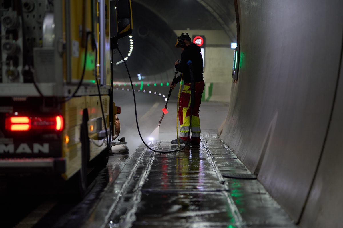 Ein Arbeiter in Warnkleidung reinigt mit einem Hochdruckreiniger eine Fahrbahn neben einem gelben Fahrzeug in einem Tunnel.