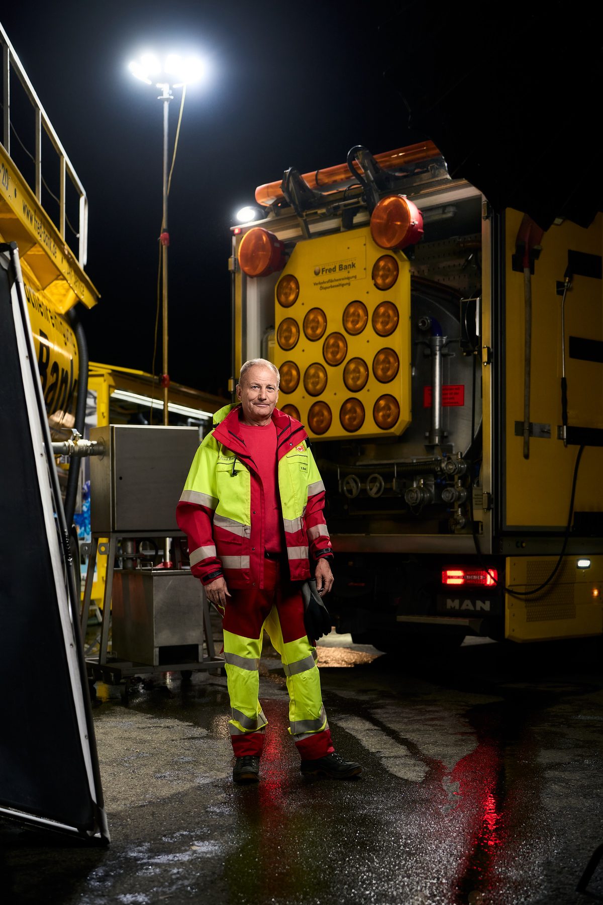 Ein Mann in roter und gelber Arbeitskleidung steht vor einem gelben Fahrzeug mit Warnleuchten, im Hintergrund ein heller Scheinwerfer.