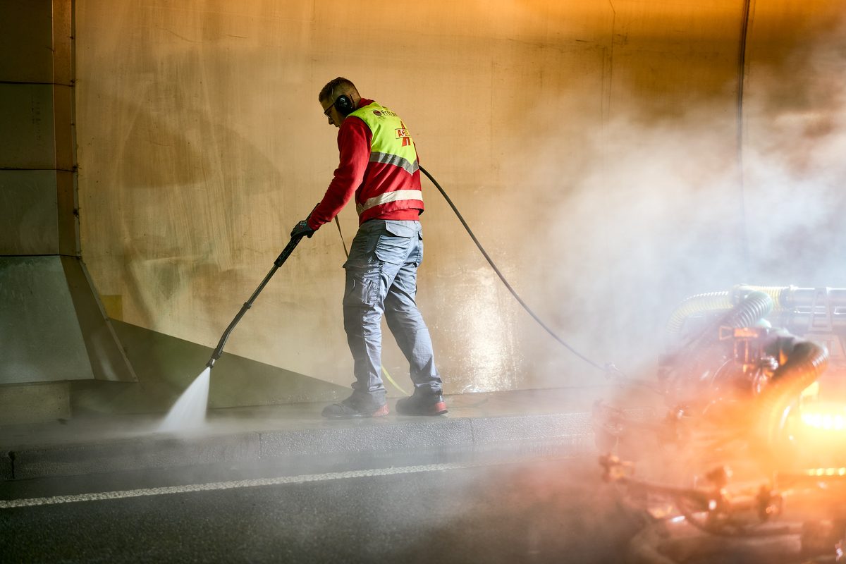 Ein Arbeiter in Warnweste und Gehörschutz reinigt mit einem Hochdruckreiniger eine Wand in einem Tunnel.