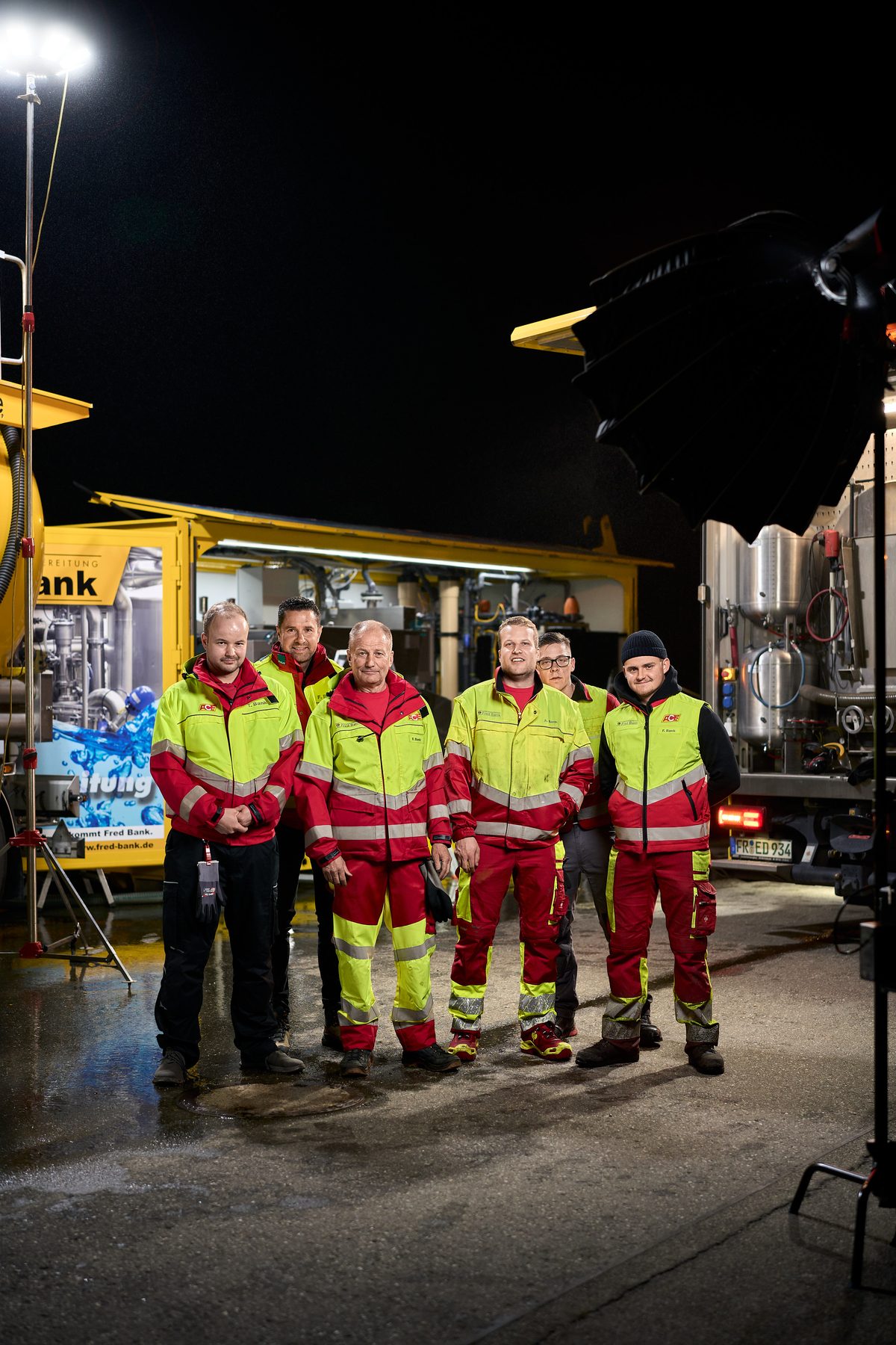 Sechs Männer in rot-gelber Arbeitskleidung stehen nachts vor gelben Industriefahrzeugen auf nassem Asphalt.