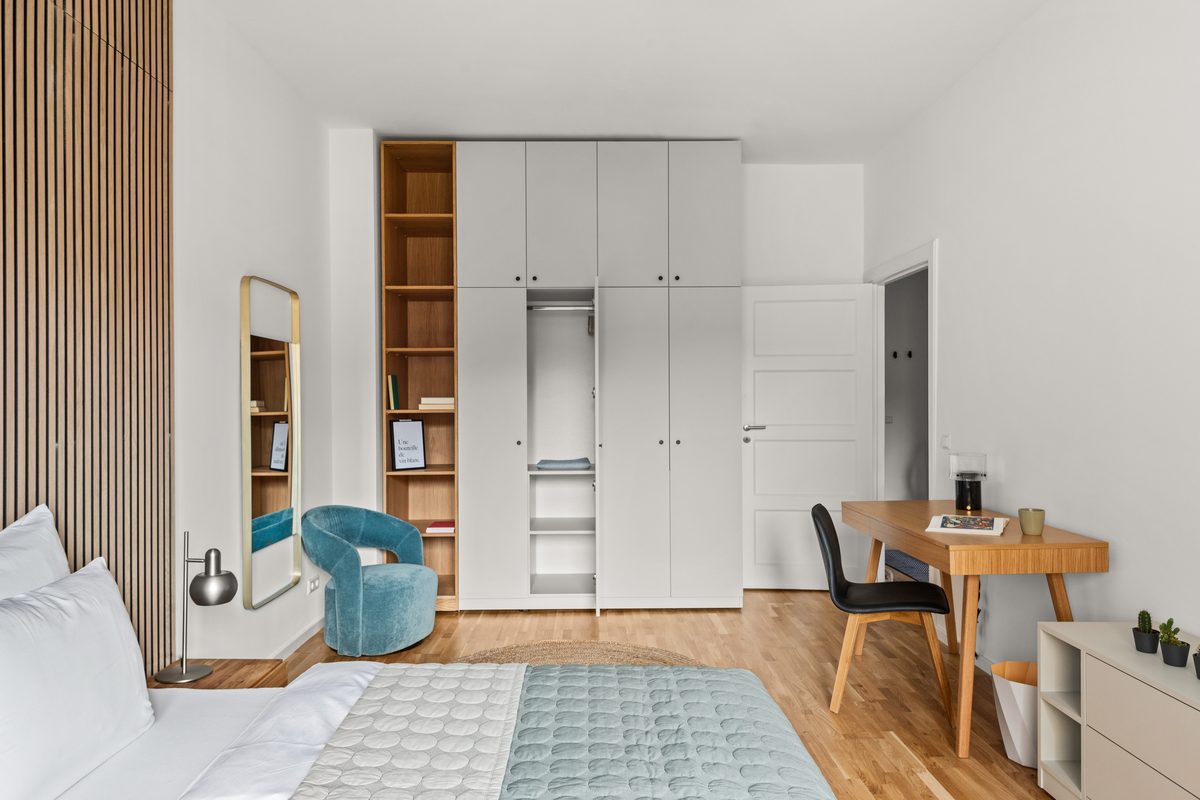Ein Schlafzimmer mit Holzboden, weißem Bett, blauem Sessel, Schrankwand, Schreibtisch und einem Spiegel.