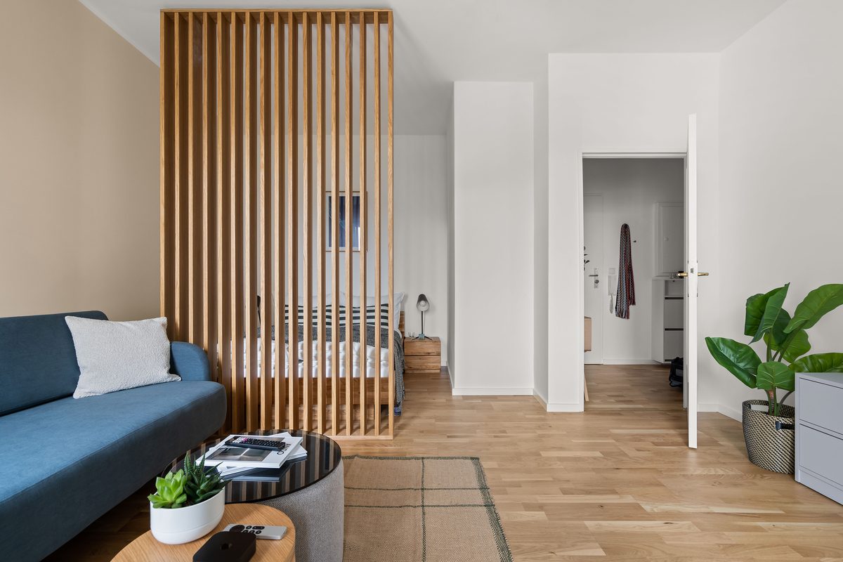 Wohnzimmer mit blauem Sofa, Couchtisch, Holzlattenwand, dahinter ein Bett und ein offener Flur mit Tür.