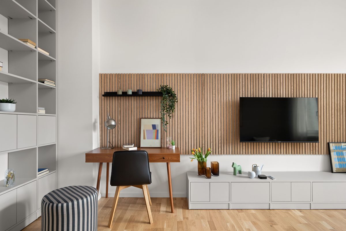 Ein modernes Wohnzimmer mit hellem Holzboden, weissen Wänden, einem Schreibtisch und einem Fernseher vor einer Holzlamellenwand.