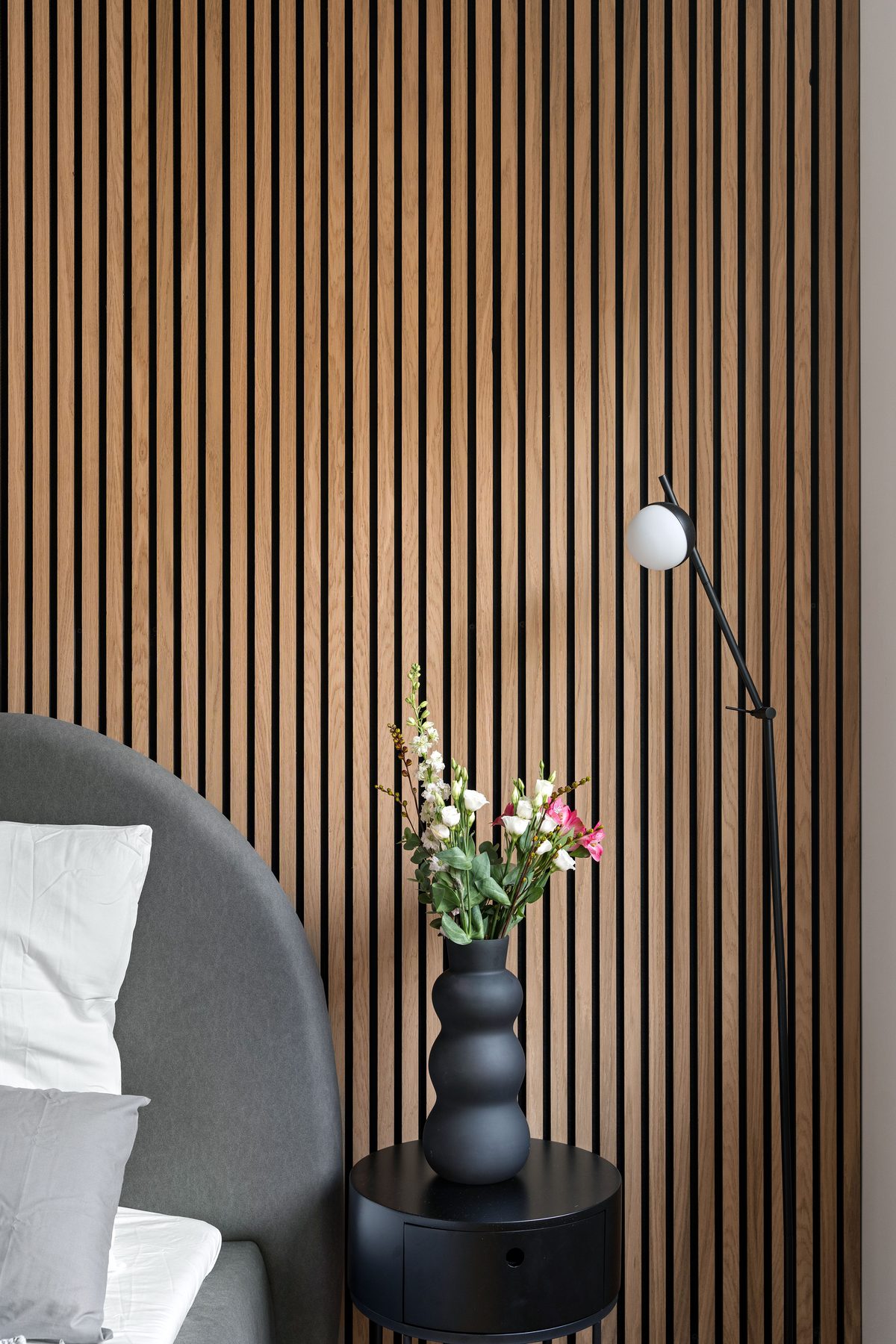 Ein Schlafzimmer mit Holzwand, einem grauen Kopfteil, einem schwarzen Nachttisch mit Vase und Blumen sowie einer Stehlampe.