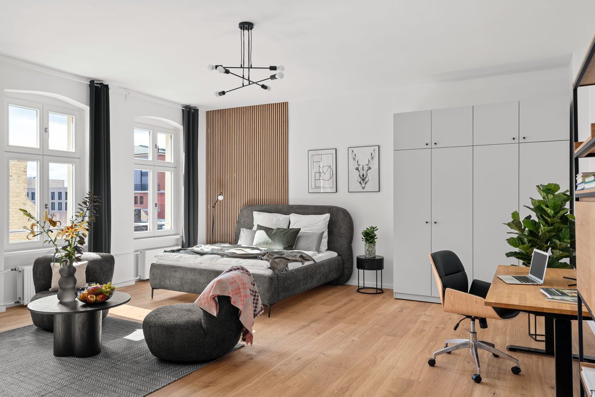 Ein Schlafzimmer mit Holzboden, einem grauen Bett, einem Schreibtisch mit Laptop und zwei Fenstern mit dunklen Vorhängen.