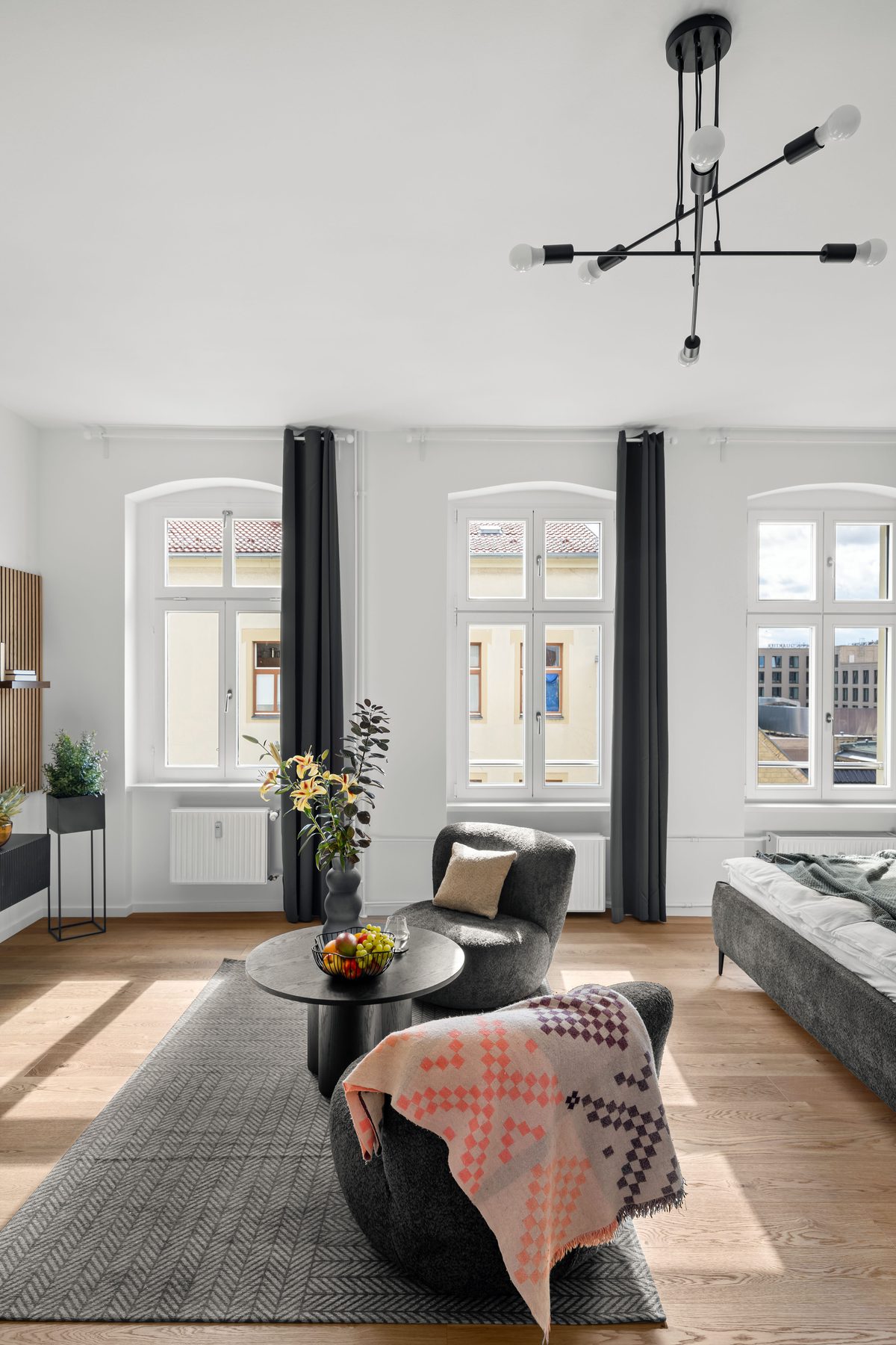 Ein helles Wohnzimmer mit Holzboden, drei Fenstern, einem grauen Teppich, zwei Sesseln, einem Tisch und einem Bett.