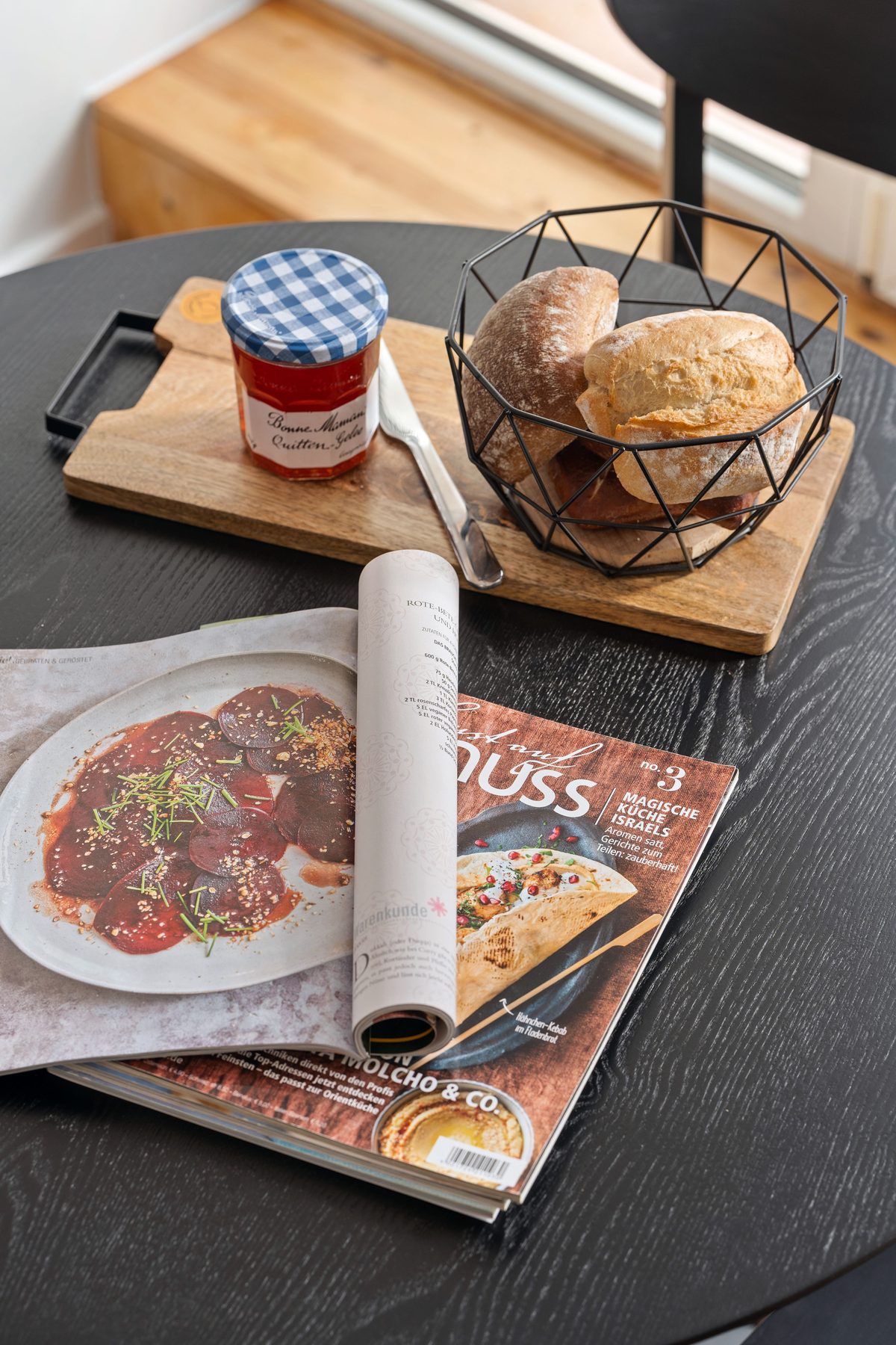 Ein Frühstückstablett mit Brötchen, Marmelade und Messer auf einem schwarzen Tisch neben einer Zeitschrift.