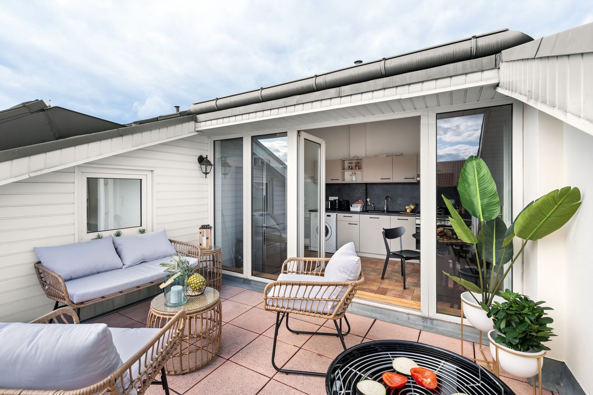 Eine Dachterrasse mit Rattanmöbeln, einem Grill und Blick auf eine offene Küche unter bewölktem Himmel.