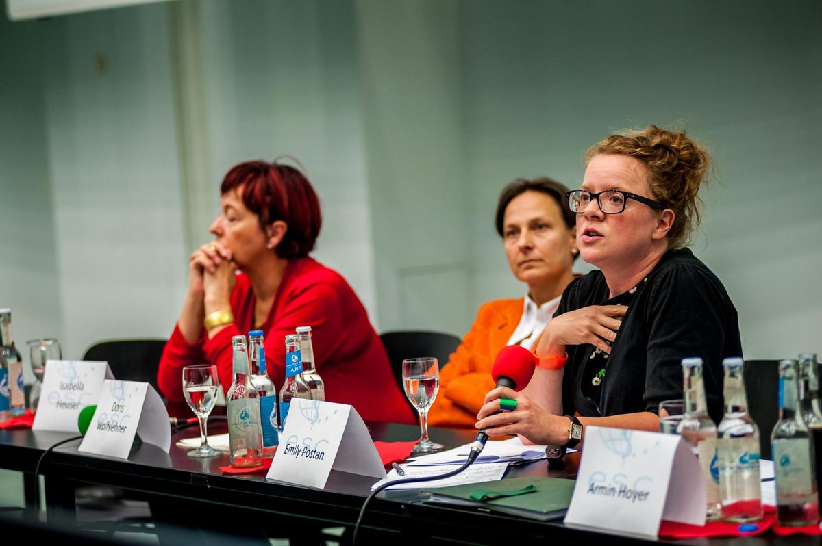 Drei Frauen sitzen an einem Tisch mit Mikrofonen, Wasserflaschen und Namensschildern.