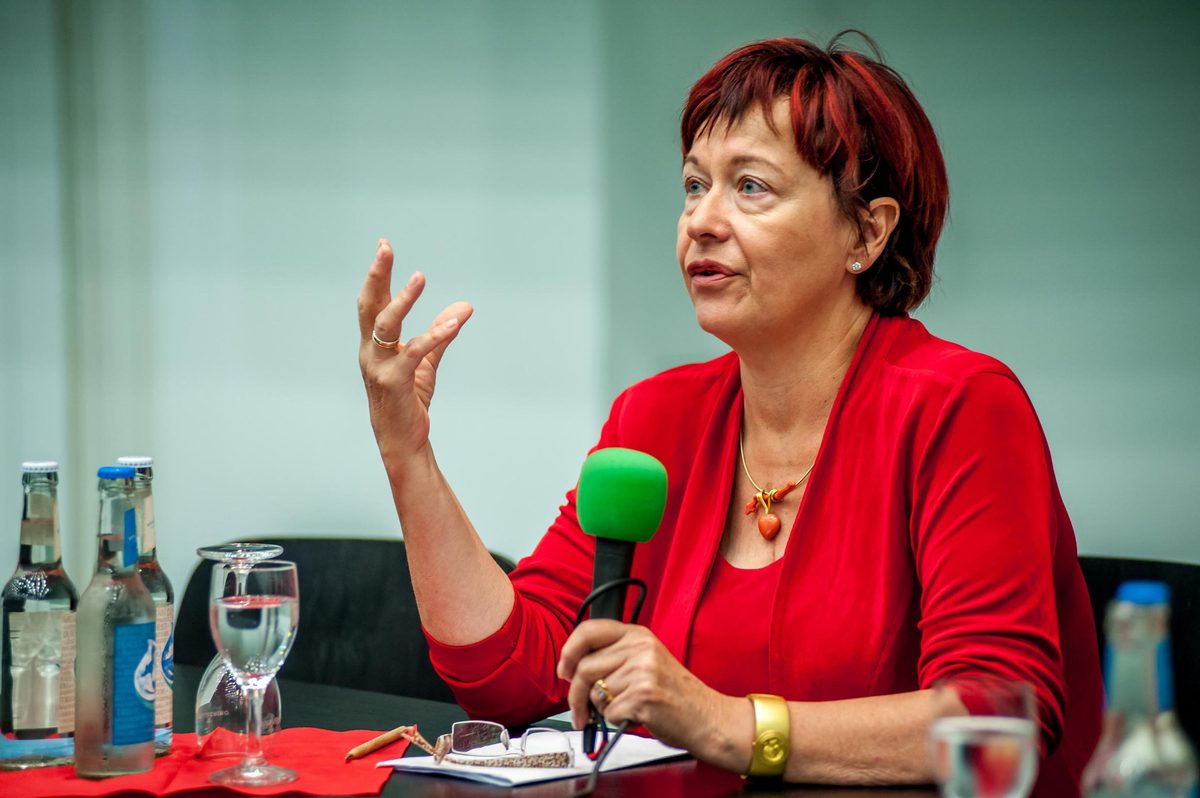 Eine Frau mit roten Haaren und roter Kleidung spricht in ein Mikrofon, gestikuliert mit der Hand.