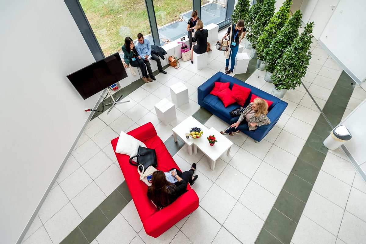 Mehrere Personen sitzen und stehen in einem modernen Raum mit roten und blauen Sofas, Tischen und Pflanzen.