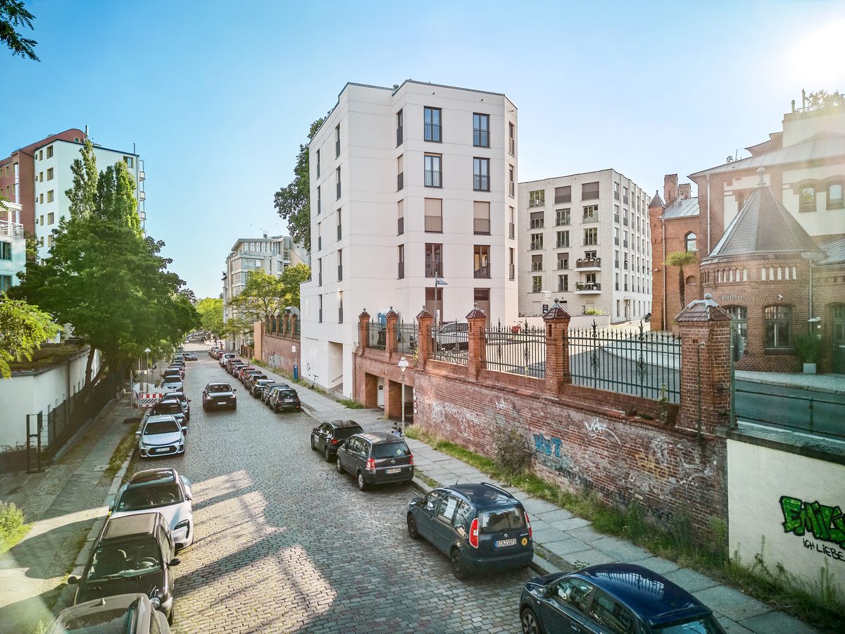 Gepflasterte Straße mit parkenden Autos, links Bäume, rechts eine Ziegelmauer und ein weißes Gebäude unter blauem Himmel.
