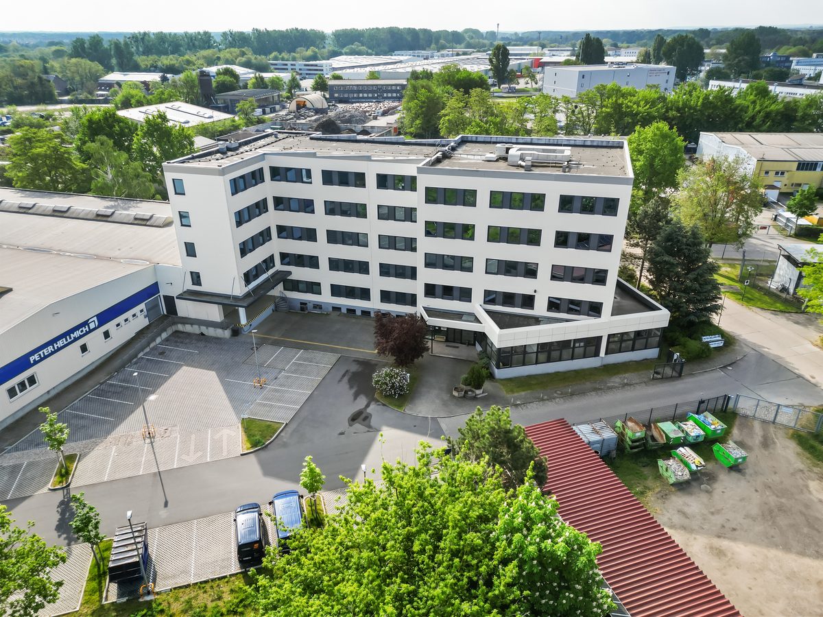 Luftaufnahme eines weißen Bürogebäudes mit Parkplatz und umliegenden Industriegebäuden und Bäumen.