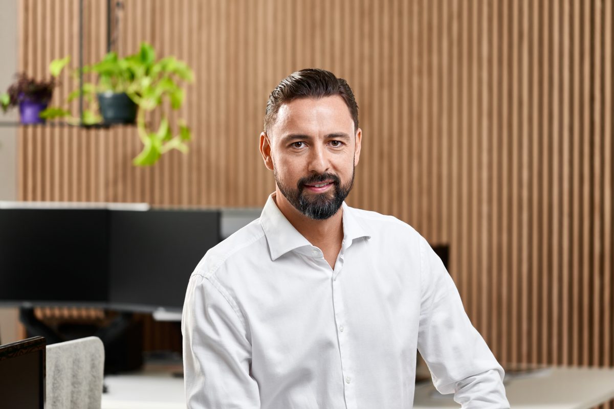 Ein Mann mit Bart und weißem Hemd lächelt in einem Büro mit Holzlamellenwand und Pflanzen im Hintergrund.