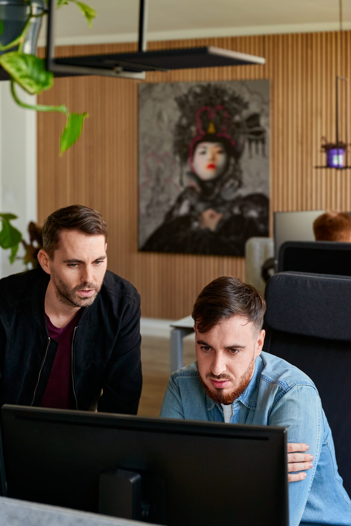 Zwei Männer schauen konzentriert auf einen schwarzen Computermonitor, im Hintergrund ein Bild einer Frau mit Kopfschmuck.
