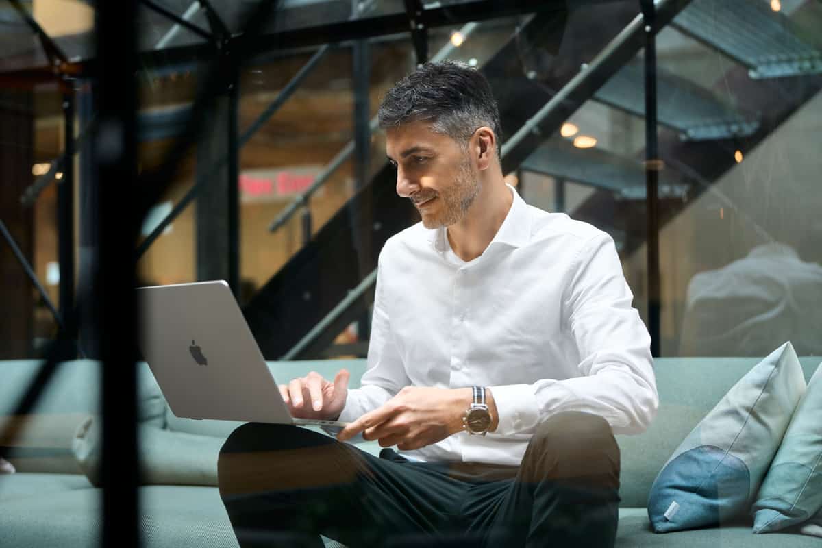 Giuseppe Conforti sitzt mit Laptop auf einem Sofa, industrielle Stahltreppe im Hintergrund.