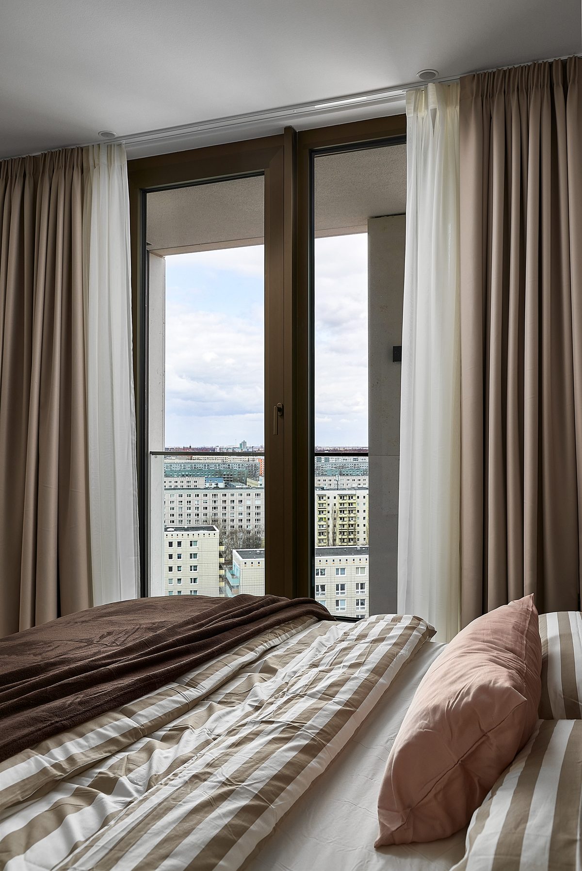 Ein Bett mit gestreifter Bettwäsche und brauner Decke vor einem Fenster mit Blick auf eine Stadtlandschaft.