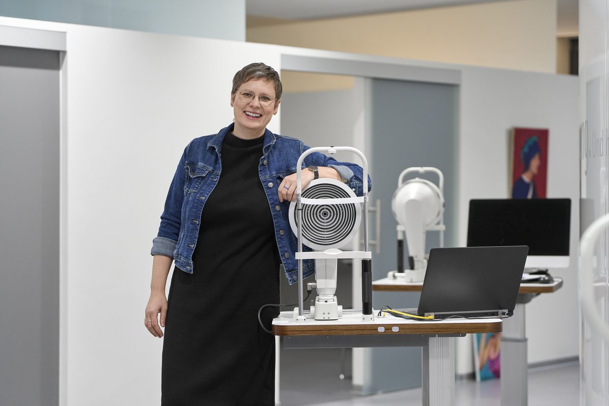 Eine lächelnde Frau mit Brille und Jeansjacke lehnt an einem weißen Augenmessgerät neben einem Laptop.