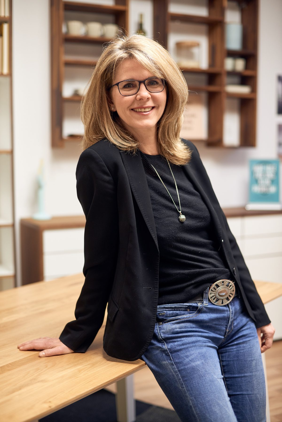 Holzconnection: Eine lächelnde Frau mit Brille, schwarzem Blazer und Jeans sitzt auf einem Holztisch vor einem Regal