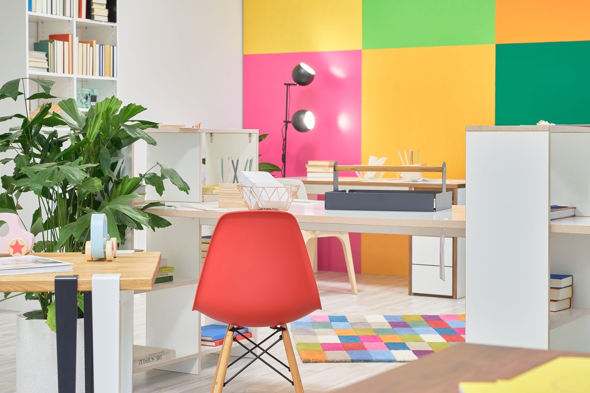 Holzconnection: Ein farbenfrohes Büro mit Schreibtischen, einem roten Stuhl, Pflanzen und einem Bücherregal vor einer bunten Wand