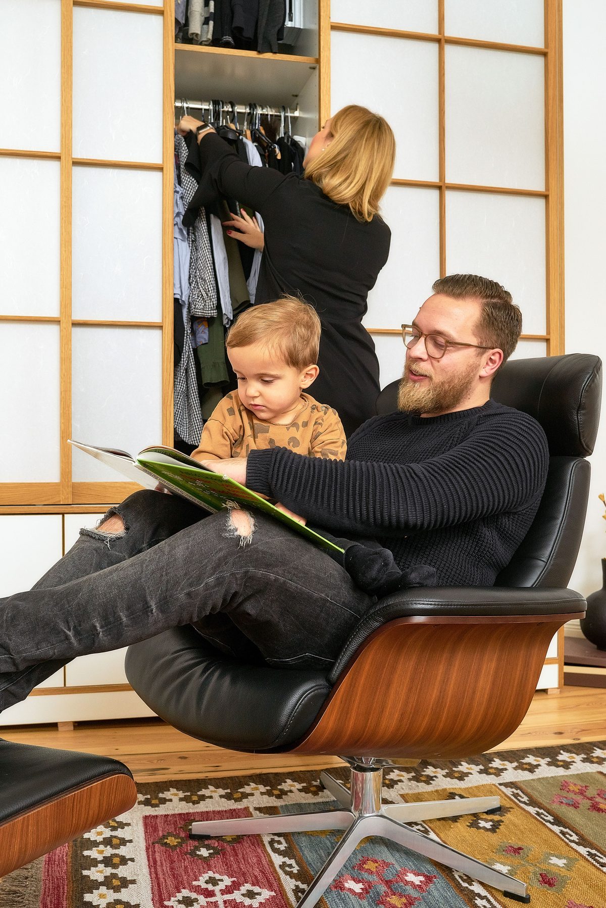 Holzconnection: Ein Mann und ein Kind lesen ein Buch in einem Sessel, während eine Frau Kleidung aus einem Schrank nimmt