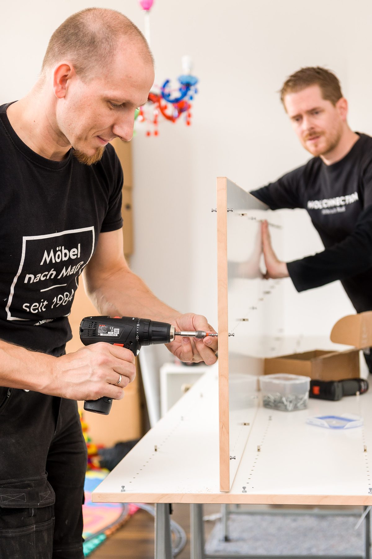 Holzconnection: Zwei Männer montieren ein weißes Regal mit Holzkanten, einer schraubt, der andere hält ein Brett fest