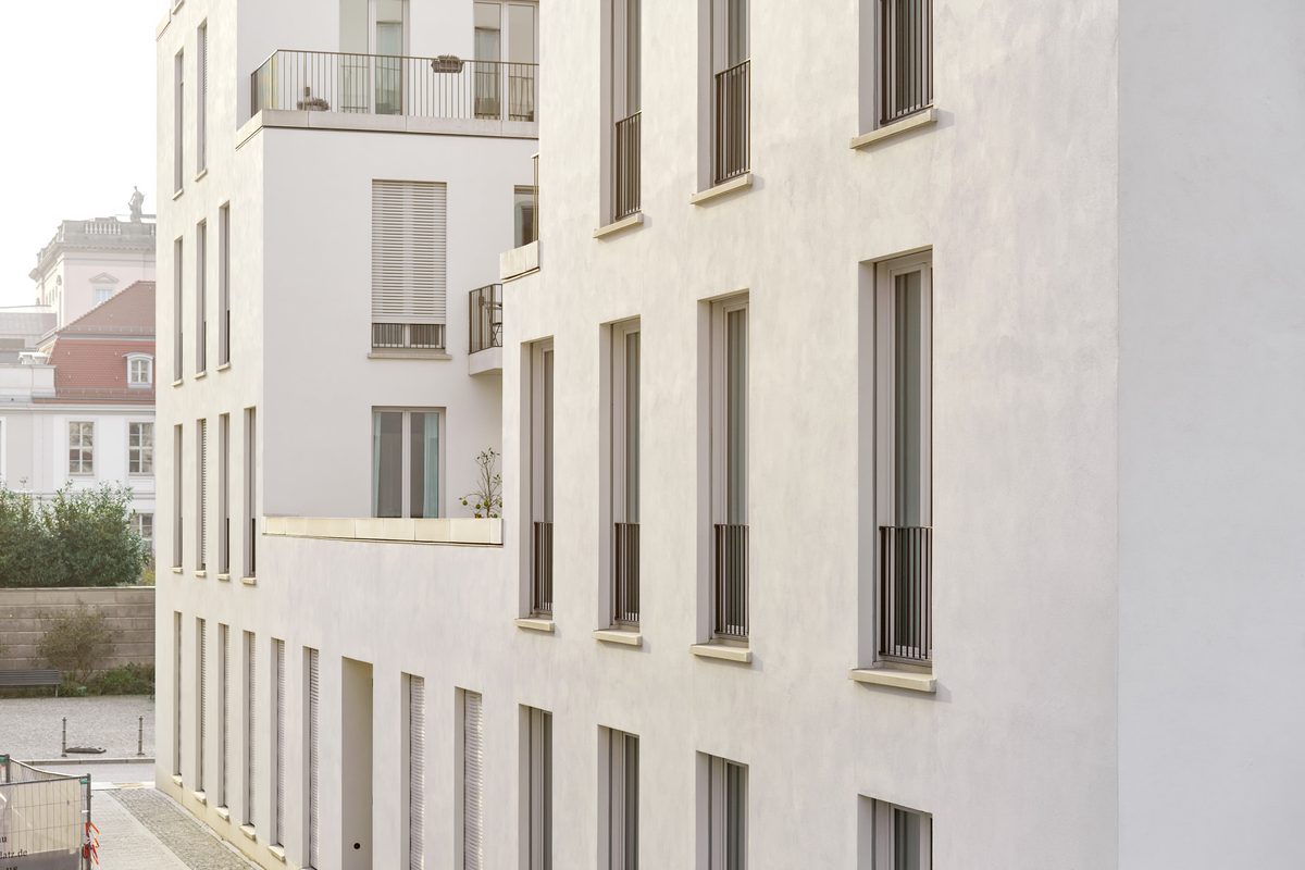 Fassade eines modernen, weißen Gebäudes mit vielen Fenstern und Balkonen, im Hintergrund ein weiteres Gebäude mit rotem Dach.
