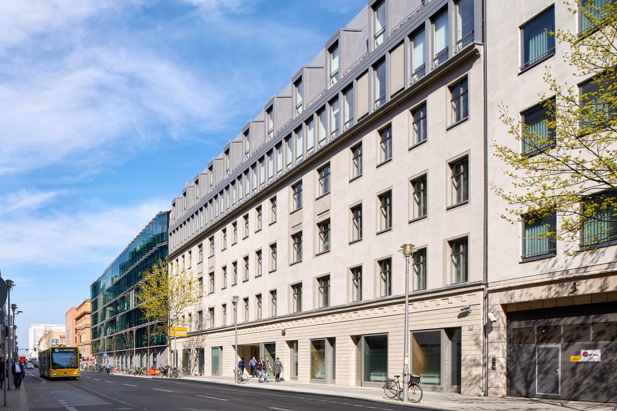 Eine Straße mit einem gelben Bus, Fahrrädern und Fußgängern vor modernen Gebäuden mit Glas- und Putzfassaden.