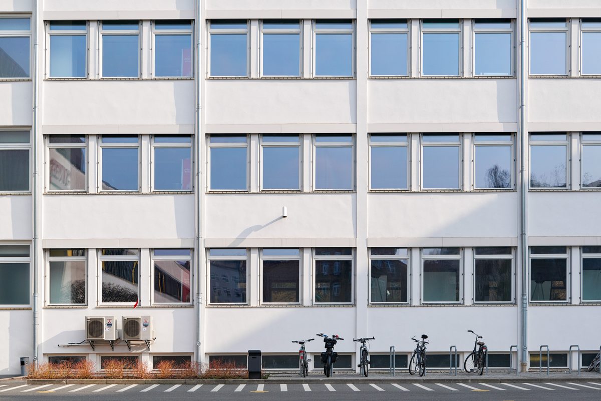 Fassade eines weißen Bürogebäudes mit vielen Fenstern, Klimaanlagen, Büschen und Fahrrädern vor einem Parkplatz.