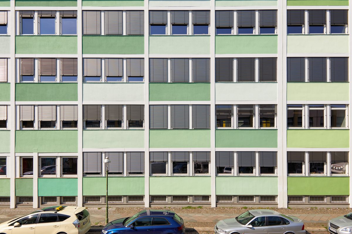 Fassade eines mehrstöckigen Gebäudes mit grünen und hellgrünen Paneelen, vielen Fenstern mit Jalousien und parkenden Autos davor.