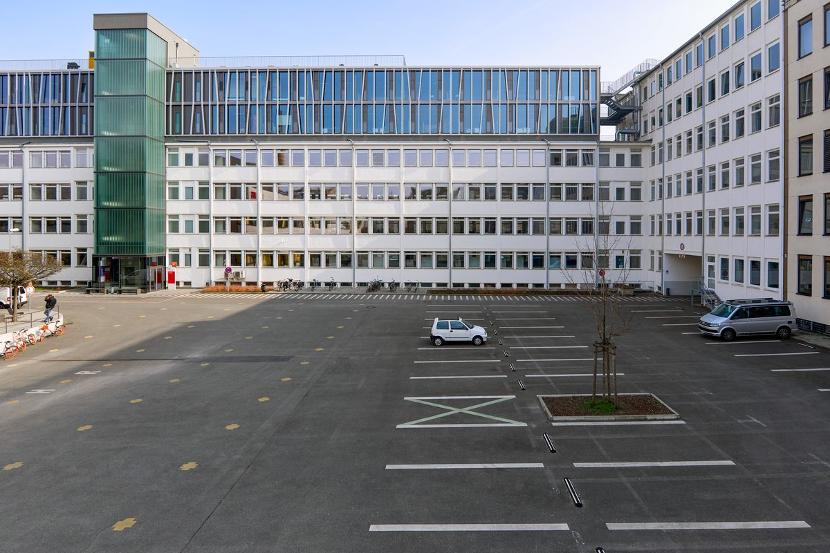 Ein Innenhof mit Parkplätzen vor einem großen weißen Gebäude mit vielen Fenstern und einem modernen Glasanbau.
