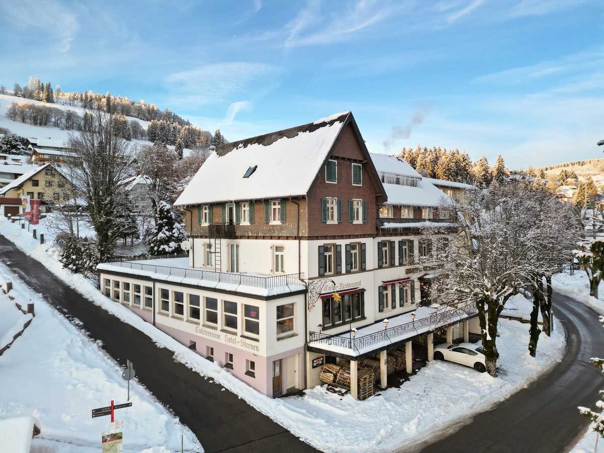 Ein großes, schneebedecktes Hotelgebäude mit Holzdach und grünen Fensterläden steht an einer Straße in einer Winterlandschaft.