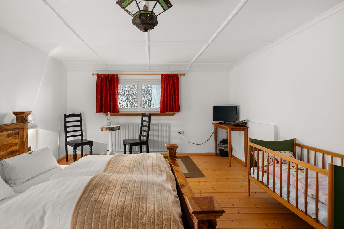 Ein Schlafzimmer mit Holzbett, Babybett, zwei Stühlen, einem Fernseher und einem Fenster mit roten Vorhängen.