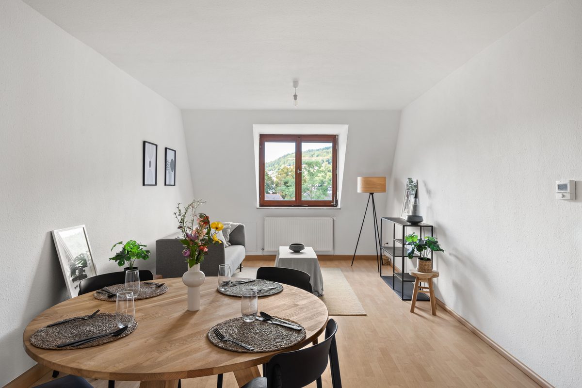 Ein heller Wohnraum mit Holzboden, Esstisch, Sofa und einem Fenster mit Blick auf bewaldete Hügel.
