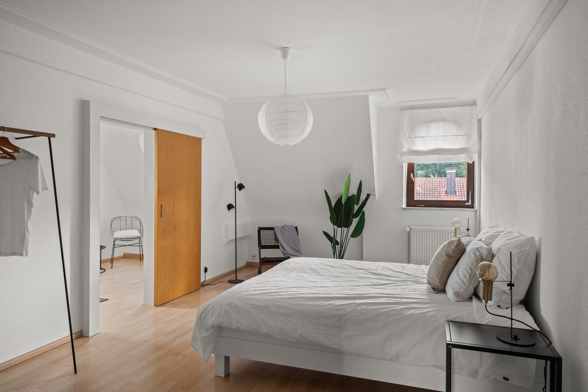 Ein helles Schlafzimmer mit weißem Bett, Nachttisch, Stehlampe, Fenster und einer offenen Holztür zu einem weiteren Raum.