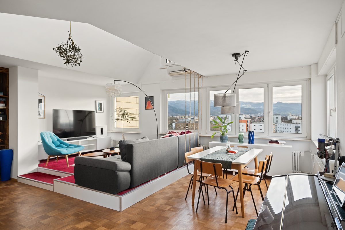 Ein Wohnzimmer mit Parkettboden, einem grauen Sofa, einem Esstisch und Blick auf eine Stadt mit Bergen im Hintergrund.