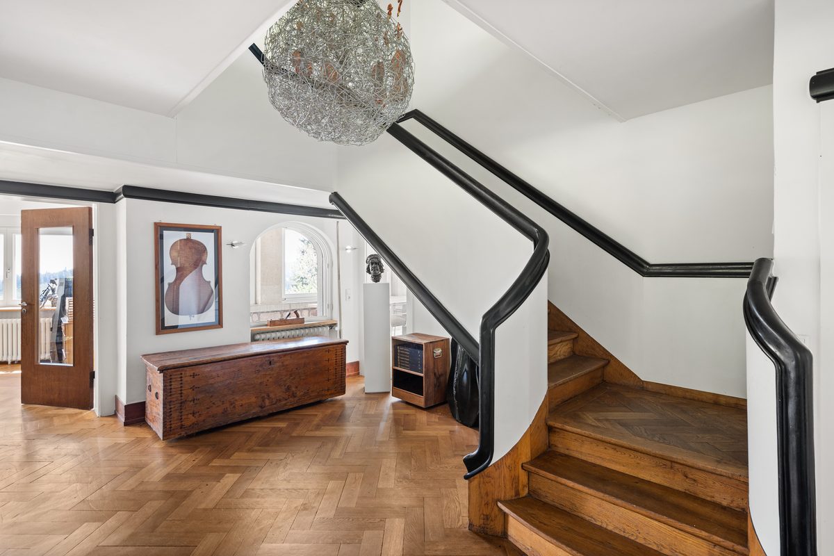 Ein Treppenhaus mit Holztreppe, Parkettboden, weiß gestrichenen Wänden und einem großen Holzkasten unter einem Fenster.