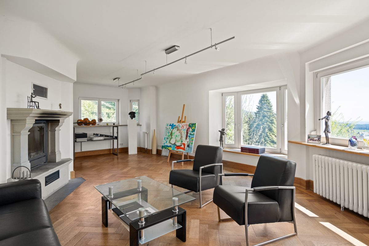 Heller Wohnraum mit Fischgrätparkett, Kamin, zwei schwarzen Sesseln, Glastisch und Fenstern mit Blick ins Grüne.