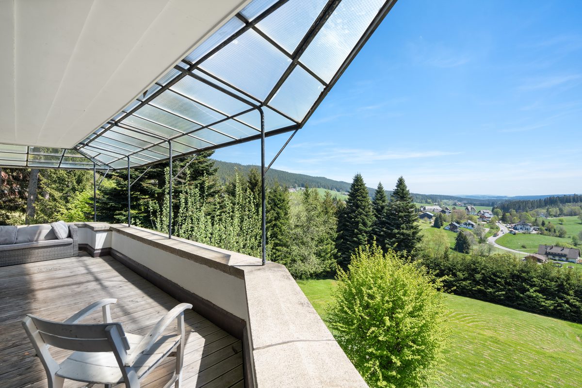 Blick von einer Terrasse mit Glasdach auf eine bewaldete Hügellandschaft mit Häusern und einem Tal unter blauem Himmel.